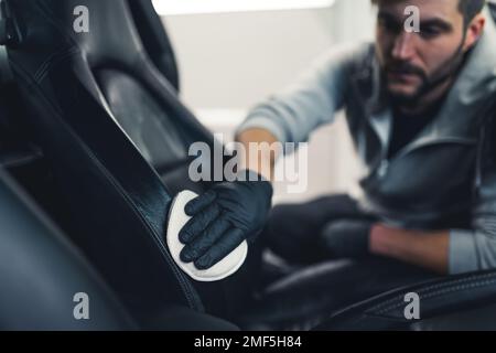 Ein weißer Mann mit schwarzen Handschuhen, der sich in das Auto lehnt, Lederbezüge für Autositze imprägniert. Schutzbeschichtung. Professionelle Fahrzeugdetails. Konzentrieren Sie sich auf den Vordergrund. Horizontale Indoor-Aufnahme. Hochwertiges Foto Stockfoto