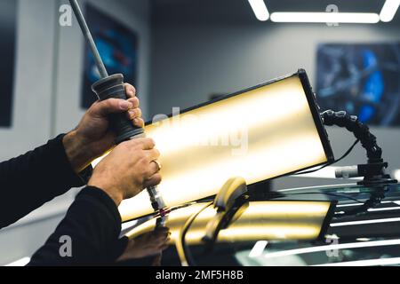 Ein unbekannter Mann entfernt Dellen im Auto mit professioneller Ausrüstung und Lampe in einer Garage. Professionelle Autoreparatur. Autoteile. Horizontale Indoor-Aufnahme. Hochwertiges Foto Stockfoto