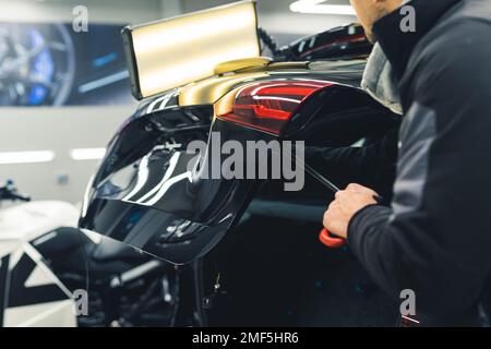 Rückansicht eines Mannes, der Vertiefungen im Fahrzeugheck mit einer hellen Lampe entfernt. Autoreparatur und Detaillierung in der professionellen Garage. Horizontaler Innenschuss. Hochwertiges Foto Stockfoto