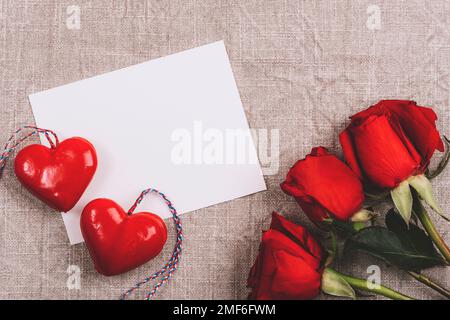 Unbeschriftete Karte, zwei Herzen und rote Rosen auf neutralem Textilhintergrund. Valentinstag-Konzept. Draufsicht, flaches Lay, Modell. Stockfoto