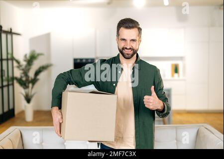 Glücklicher attraktiver, eleganter Kaukasier, zu Hause stehen, einen großen Karton in den Händen halten, ein Paket aus dem Online-Shop erhalten, sich auf das Auspacken vorbereiten, in die Kamera schauen, lächeln, Daumen-hoch-Geste Stockfoto