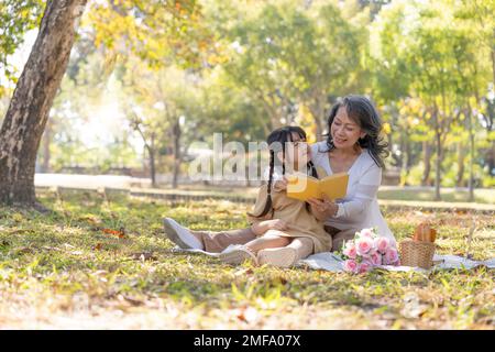 Fröhliche asiatische Großmutter, die ihrer bezaubernden Enkelin eine Geschichte erzählt, während sie am Wochenende ein Picknick im wunderschönen Park macht Stockfoto