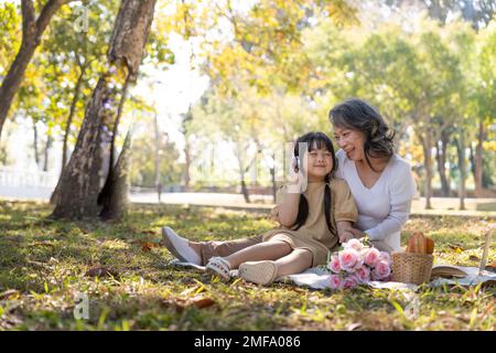 Fröhliche asiatische Großmutter, die mit ihrer reizenden Enkelin zusammen im Park picknickt. Freizeit- und Familienkonzept