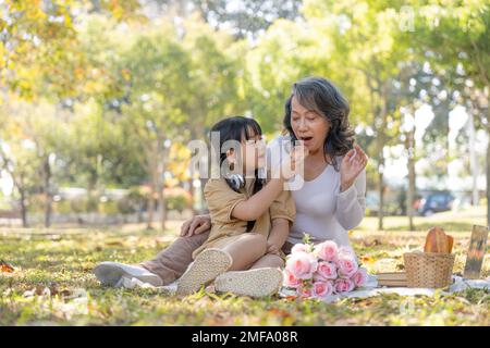 Fröhliche asiatische Großmutter, die mit ihrer reizenden Enkelin zusammen im Park picknickt. Freizeit- und Familienkonzept Stockfoto