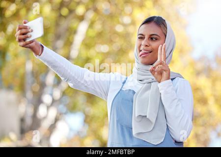 Selfie, Frieden und islamische Frau im Freien, dummes Gesicht oder selbstbewusst mit lässiger Kleidung, Glück oder Freude. Muslimische Frau, Frau in der Natur oder Smartphone Stockfoto