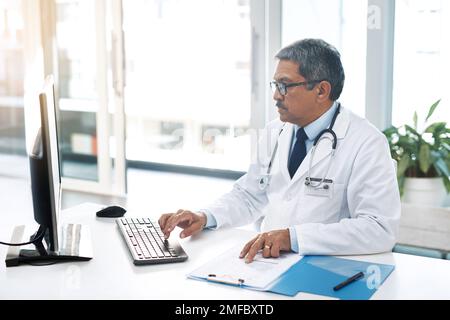 So wie es aussieht, ist alles in Ordnung. Ein fokussierter, reifer Arzt, der tagsüber an einem Computer arbeitet, während er in seinem Büro sitzt. Stockfoto