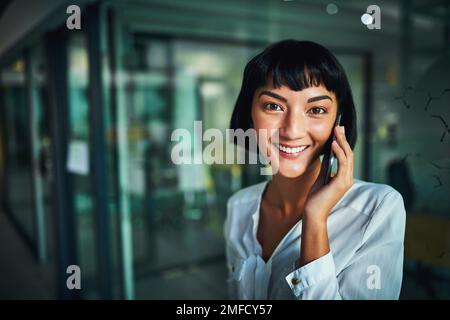 Ich stehe zur Verfügung, um über Geschäfte zu sprechen. Eine junge Geschäftsfrau, die ein Mobiltelefon in einem modernen Büro benutzt. Stockfoto