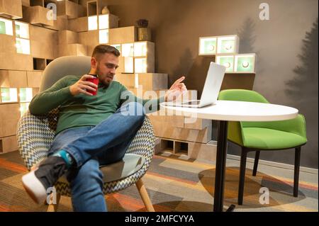 Ein weißer Mann mit Bart sitzt in einem bequemen Stuhl, trinkt Kaffee und arbeitet an einem Laptop. Freiberufler. Stockfoto