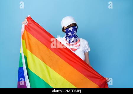 Eine weiße junge Frau, die mit einer Regenbogenflagge posiert, isoliert auf blauem Hintergrund im Studio. Ihr Gesicht ist mit feuchten usa bedeckt und trägt ein weißes T-Shirt und eine weiße Mütze Stockfoto