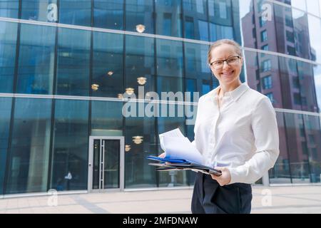 Porträt von Geschäftsfrau mit Geschäftsunterlagen, Ordner mit Akten, Frau, die draußen in weißem Hemd und schwarzer Hose mit Brille vor die Kamera schaut, Business Center oder Bürogebäude im Hintergrund Stockfoto