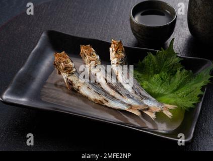 Gegrillte Uransardinen auf einem schwarzen Tisch. Eine Beilage Sake. Bild: Japanische Küche. Stockfoto