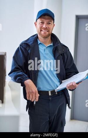 Ihr Paket wird immer pünktlich sein. Verkürztes Porträt eines gutaussehenden Lieferers, der in der Lobby mit einer Kundenbestellung wartet. Stockfoto