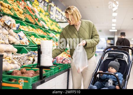 Casualy kleidete die Mutter, die Obst und Gemüse im Supermarkt kaufte, zusammen mit ihrem kleinen Jungen im Kinderwagen Stockfoto