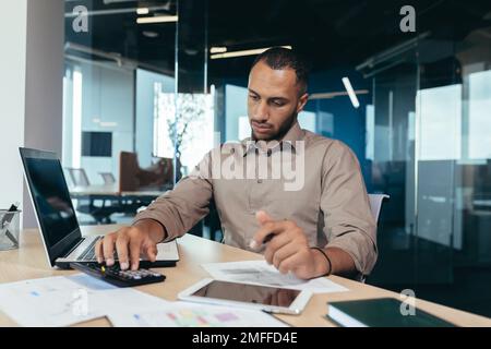 Ein junger afroamerikanischer Buchprüfer arbeitet im Büro an einem Tisch mit einem Laptop und Dokumenten. Papierarbeit, konzentrierte Berechnungen auf einem Taschenrechner. Stockfoto