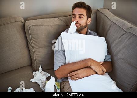 Heute gehe ich nirgendwo hin. Ein müder junger Mann, der tagsüber auf einer Couch mit einem Taschentuch in der Nase liegt und ein Kissen zu Hause hält. Stockfoto