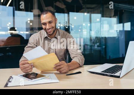 Ein glücklicher junger Mann erhielt einen Brief mit guten Nachrichten. Er sitzt im Büro an einem Tisch mit einem Laptop, hält einen Umschlag in der Hand, liest und lächelt. Stockfoto