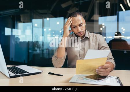 Ein aufgebrachter junger afroamerikanischer Mann liest schlechte Nachrichten aus einem Brief, den er erhielt. Er sitzt am Schreibtisch im Büro und hält seinen Kopf mit der Hand. Stockfoto