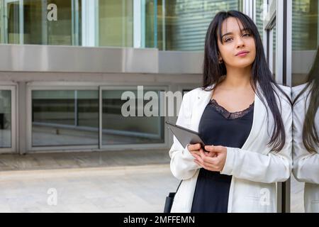 Porträt einer jungen Geschäftsfrau, das sich an einer Glaswand mit Reflexionen lehnt. Ein Lächeln befähigt eine smarte, zwanglose Frau, die das Tablet außerhalb des Büros hält. Stockfoto