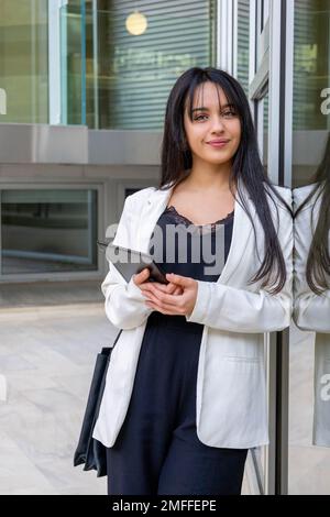 Porträt einer jungen Geschäftsfrau, das sich an einer Glaswand mit Reflexionen lehnt. Ein Lächeln befähigt eine smarte, zwanglose Frau, die das Tablet außerhalb des Büros hält. Stockfoto