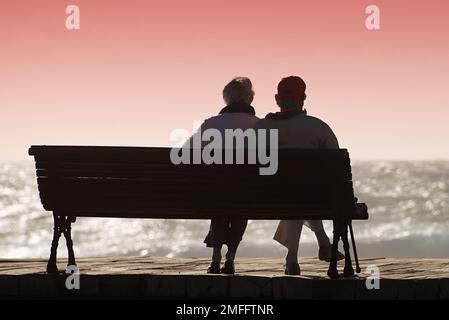 Silhouetten-Seniorenpaar, das auf den farbenfrohen Sonnenuntergang auf der Bank in der Nähe des Ozeans wartet Stockfoto