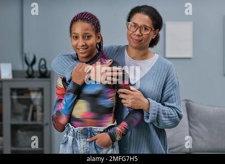 Taille hoch Porträt von schwarzer reifer Frau, die ihre Teenager-Tochter in minimalem Innenraum umarmt und beide in die Kamera schaut Stockfoto