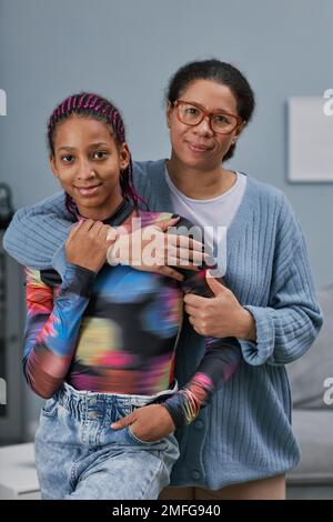 Vertikales Porträt einer reifen Mutter, die eine Teenager-Tochter umarmt, in minimalem Heimbereich, beide in die Kamera schaut Stockfoto