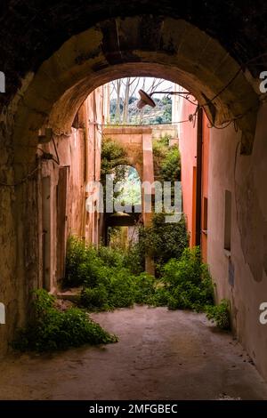 Eine kleine, zerstörte Gasse mit Torbögen, Steinmauern und überwucherten Treppen im alten Teil der Stadt Ragusa. Stockfoto