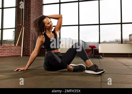 Attraktive afroamerikanische Frau in Sportbekleidung, die in die Kamera schaut und lächelt, während sie in einem Crossfit-Fitnessstudio auf dem Boden sitzt Stockfoto