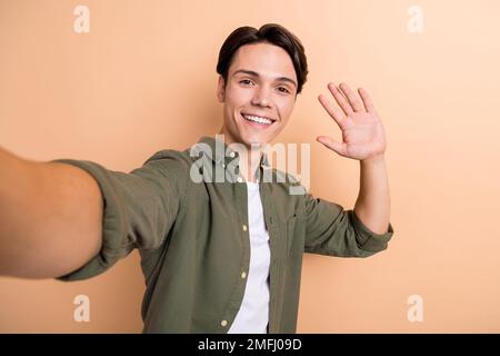 Foto eines optimistischen, gut aussehenden, coolen Typen mit braunem Haar, gekleidetem Khaki-Hemd, der Selfie mit der Hand wedelt, isoliert auf beigefarbenem Hintergrund Stockfoto