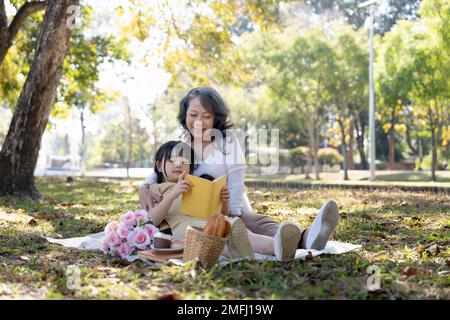 Fröhliche asiatische Großmutter, die ihrer bezaubernden Enkelin eine Geschichte erzählt, während sie am Wochenende ein Picknick im wunderschönen Park macht Stockfoto