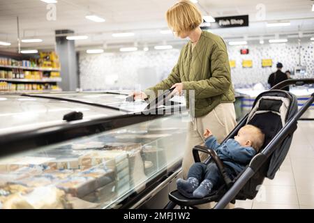 Casualy kleidete Mutter, die Tiefkühlprodukte in der Supermarkt-Abteilung auswählte, zusammen mit ihrem kleinen Jungen im Kinderwagen Stockfoto