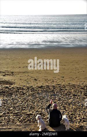 Das Bild zeigt einen Mann mit zwei Hunden, der in Coney, Sandy Beach, Porthcawl, South Wales an einer Wand saß. Er schaut mit der Flut auf das Meer hinaus. Stockfoto