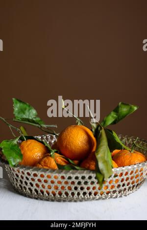 Stillleben von Mandarinen, Mandarinen, Orangenfrucht mit grünen Blättern in einem silbernen Metallkorb an der braunen Wand Stockfoto