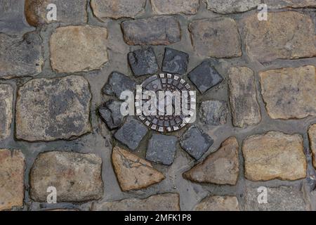 Alte Abwasserluke aus Metall in der Mitte der Straße, aus einem glatten Pflasterstein. Stockfoto