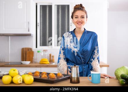 Hübsche Frau im Morgenmantel, die Kaffee trinkt und Muffins in der Küche isst Stockfoto