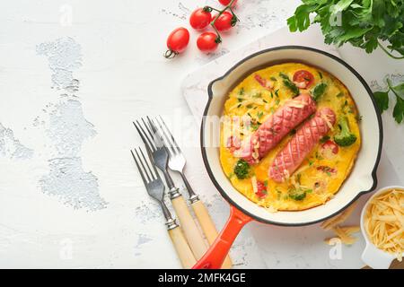 Omelette oder Frittata mit zwei Würstchen, geschmolzenem Käse, Kirschtomaten, Brokkoli und Petersilie auf roter Pfanne oder Eisentopf auf weißem, altem Betonrücken Stockfoto
