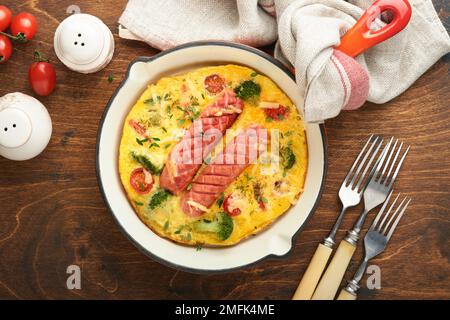 Omelette oder Frittata mit zwei Würstchen, geschmolzenem Käse, Kirschtomaten, Brokkoli und Petersilie auf roter Pfanne oder Eisenpfanne auf altem rustikalem Holzbacken Stockfoto