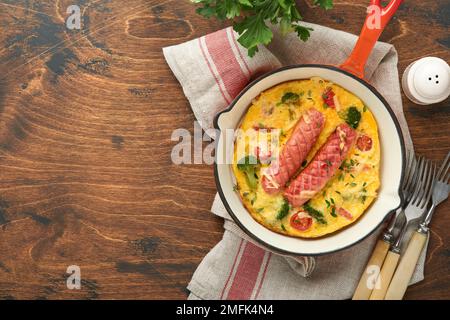 Omelette oder Frittata mit zwei Würstchen, geschmolzenem Käse, Kirschtomaten, Brokkoli und Petersilie auf roter Pfanne oder Eisenpfanne auf altem rustikalem Holzbacken Stockfoto