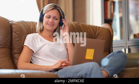 Frau mit lächelnden Kopfhörern und Blick auf den Laptop-Bildschirm, während sie auf der Couch sitzt. Stockfoto
