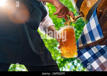 Ein bayerischer Mann in der Schürze schüttet ein großes Lagerbier aus dem Holzfass im Biergarten. Hintergrund für Oktoberfest, Folk- oder Bierfest Stockfoto