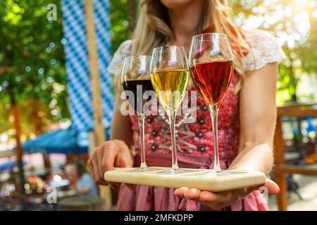 Die Kellnerin bringt verschiedene Weinsorten in Gläsern durch einen bayerischen Biergarten oder ein oktoberfest Stockfoto
