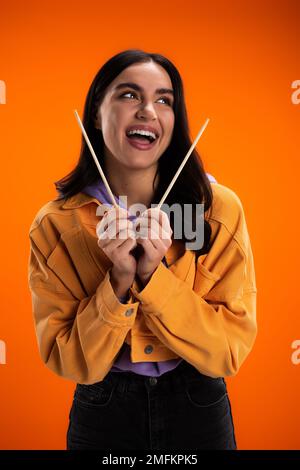 Fröhliche junge Frau, die Bambusstäbchen in der Hand hält und isoliert auf einem orangefarbenen Stockbild wegschaut Stockfoto