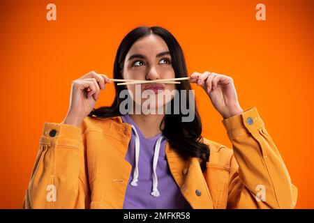 Junge Frau hält Bambusstäbchen nahe dem Mund und schmollt Lippen isoliert auf orangefarbenem Stockbild Stockfoto