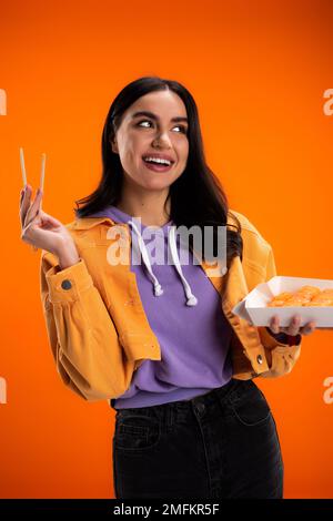 Positive junge Frau, die Essstäbchen und Sushi zum Mitnehmen hält, isoliert auf orangefarbenem Stock-Bild Stockfoto