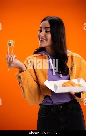Stilvolle junge Frau, die Sushi und Essstäbchen isoliert auf einem orangefarbenen Stockbild ansieht Stockfoto