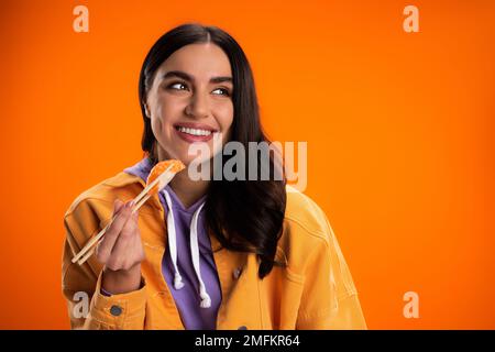 Lächelnde Frau in trendiger Jacke mit Stäbchen und frischem Sashimi isoliert auf orangefarbenem Stockbild Stockfoto