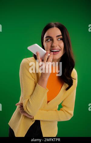 Fröhliche brünette Frau in orangefarbenem Rollkragen und gelbem Pullover, die Sprachnachricht auf Smartphone isoliert auf grünem Stockbild aufnimmt Stockfoto
