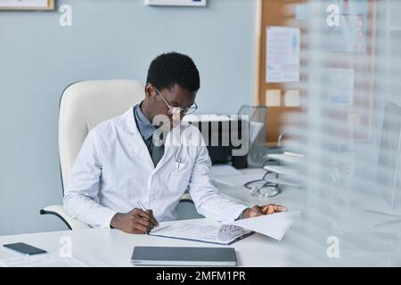 Porträt eines jungen schwarzen Arztes, der während der Arbeit in einer Privatklinik am Schreibtisch in der Praxis Papiere ausfüllt Stockfoto