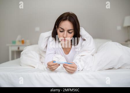 Verwirrte junge Frau, die ein elektronisches Thermometer in ihren Händen sieht Stockfoto