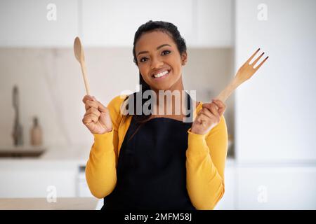 Eine fröhliche, afroamerikanische Chefköchin mit Holzlöffel und Gabel in der Schürze, die Essen kocht Stockfoto